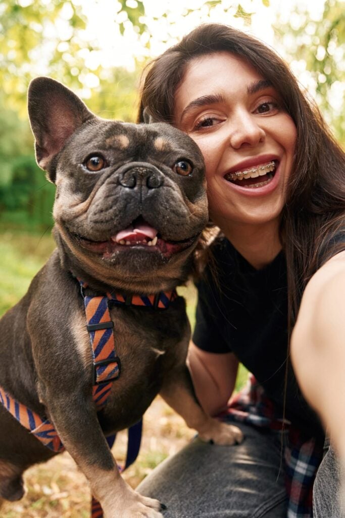 Woman with braces smiling next to her happy French bulldog outdoors.